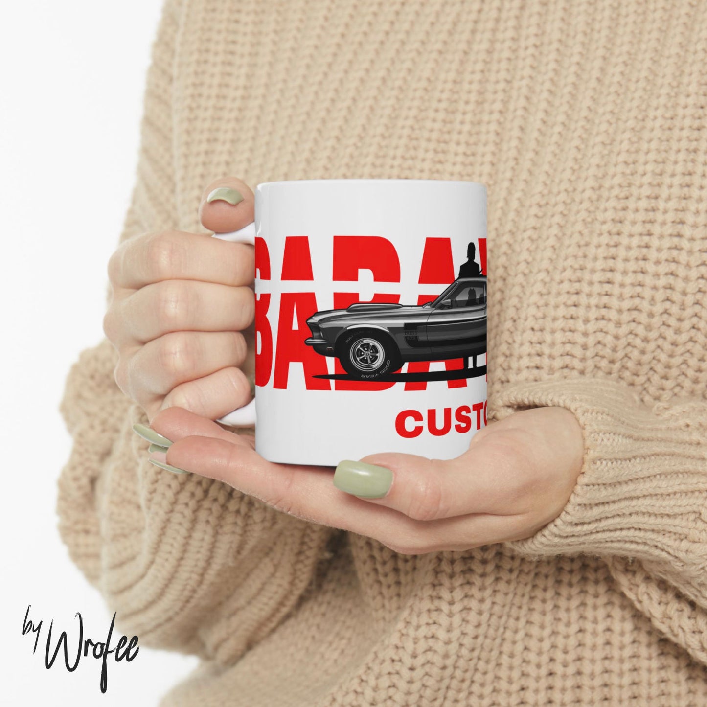 Person holding a mug with a car design and text, wearing a beige sweater.