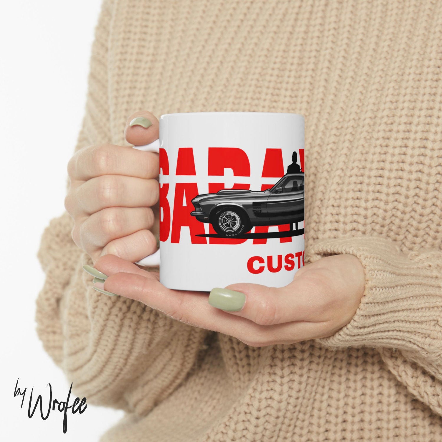 Person holding a mug with a car design and text, wearing a beige sweater.
